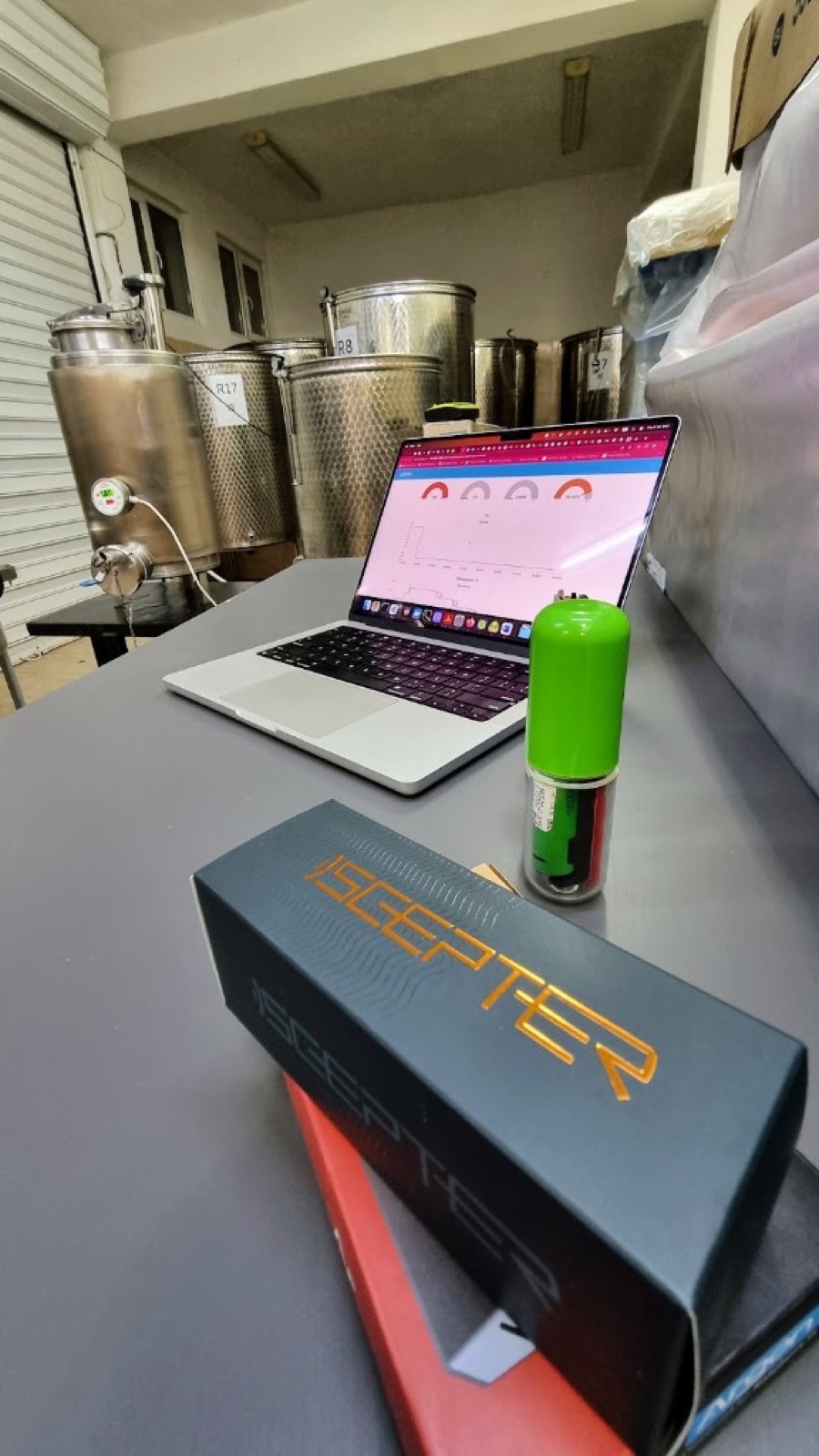 Workshop bench with a laptop showing the SCEPTER dashboard, a green float device and branded SCEPTER boxes, steel tanks behind